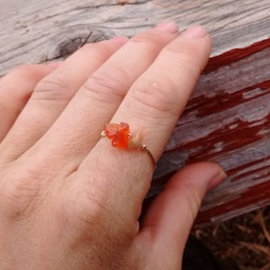 May include: A delicate gold ring with a cluster of orange carnelian stones. The ring is worn on a hand resting on a weathered wooden surface.