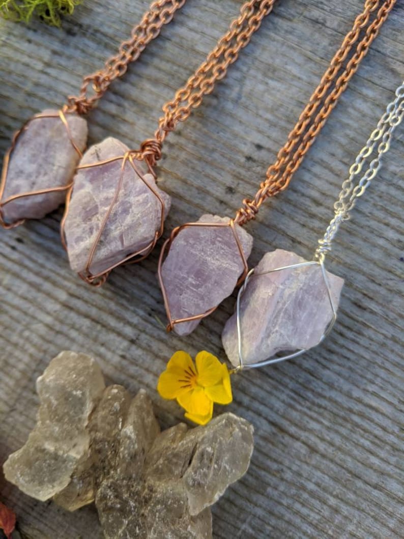 May include: Three necklaces with pink crystal pendants wrapped in copper wire. The necklaces are on a wooden surface with a yellow flower.