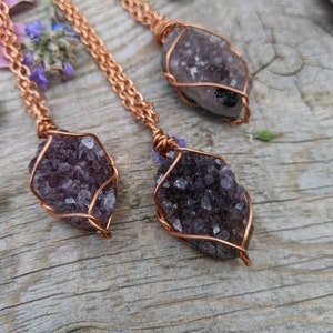 May include: Three amethyst crystal pendants wrapped in copper wire, hanging from copper chains. The pendants are on a wooden surface.