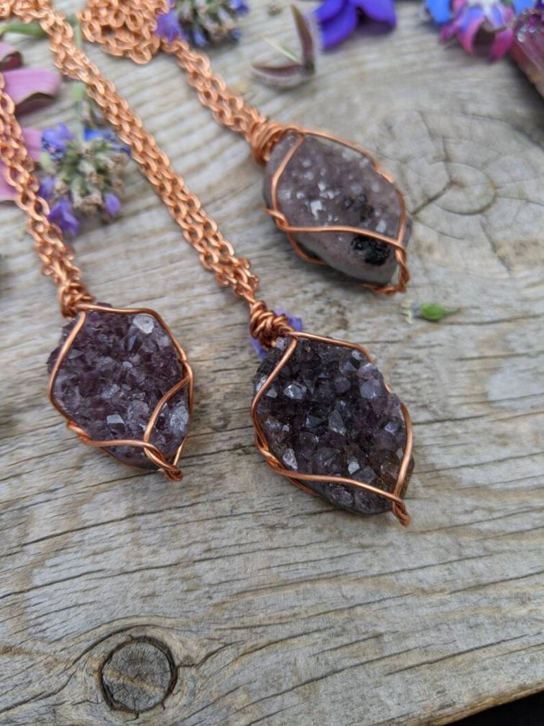 May include: Three amethyst crystal pendants wrapped in copper wire, hanging from copper chains. The pendants are on a wooden surface.