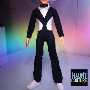 May include: A doll wearing a black and white jumpsuit with a bow tie and white sneakers. The doll has a disco ball above its head. The text "HAUNT COUTURE" is in the lower right corner.