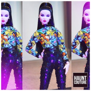 Pu&ograve; includere: Una bambola di moda che indossa un top corto nero e blu con una stampa Pokemon e pantaloni neri di paillettes. La bambola ha lunghi capelli neri e indossa un choker nero. Lo sfondo &egrave; un gradiente viola e rosa. Il testo "HAUNT COUTURE" &egrave; visibile nell'angolo in basso a destra dell'immagine.