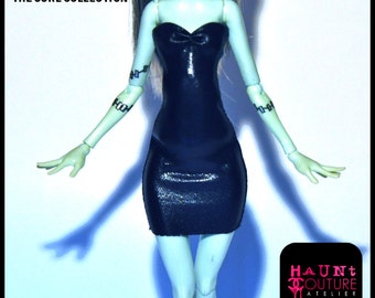 May include: A green-skinned doll wearing a black strapless dress with a bow at the neckline. The doll has black stitching on her arms and a black choker necklace. The doll is standing on a white background. The text "ESSENTIALS THE CORE COLLECTION" is at the top of the image. The text "HAUNT COUTURE ATELIER" is at the bottom of the image.