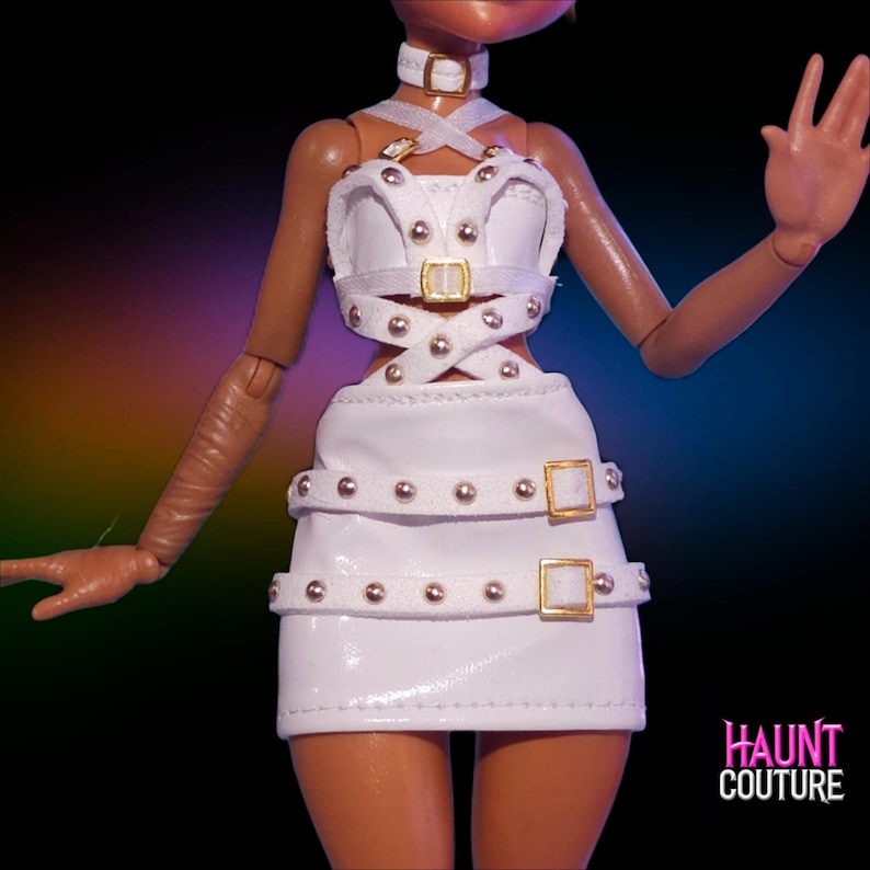 May include: A white doll-sized outfit with gold-colored buckles and studded accents. The outfit includes a top, skirt, and choker. The text "HAUNT COUTURE" is visible in the lower right corner. The outfit is designed with a unique, edgy style.