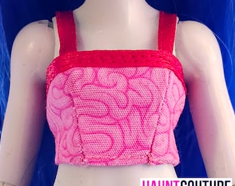 Puede incluir: Un top corto rosa y rojo con un patrón único. El top presenta un escote cuadrado y tirantes en un color rojo vibrante. La tela rosa tiene un diseño texturizado. El texto "HAUNT COUTURE" es visible en la parte inferior.