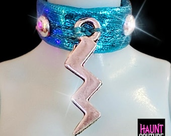 Puede incluir: Un collar gargantilla turquesa con un colgante de rayo plateado. La gargantilla tiene un acabado texturizado e iridiscente y está adornada con dos tachuelas redondas de color plateado. El logotipo de Haunt Couture está en la esquina inferior derecha.