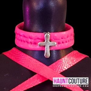 May include: A pink leather choker with a silver cross pendant. The choker has stitching detail and is worn on a mannequin.
