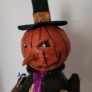 以下が含まれることがあります： 黒いシルクハットと紫のリボンをつけたカボチャのハロウィン飾り。カボチャは大きく曲がった鼻といたずらっぽい笑顔の彫刻が施されています。飾り物は小さな骸骨を手に持っています。