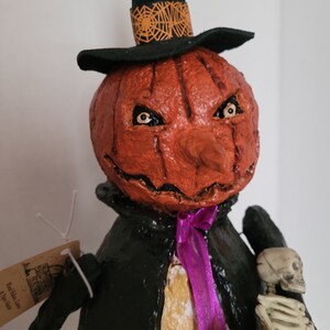 May include: A handmade paper mache pumpkin with a black top hat and a purple ribbon. The pumpkin has a scary face with orange and brown paint. The pumpkin is holding a small skeleton figure.