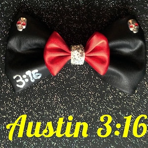 May include: A black leather bow with a red satin center and silver glitter trim. The bow has two silver skull embellishments and the text "3:16" written on one side. The text "Austin 3:16" is written in yellow on a black sparkly background.