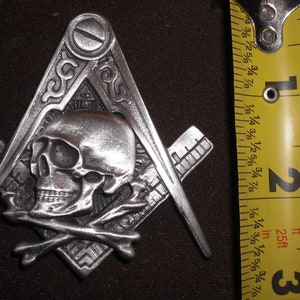 May include: A silver-toned metal pendant in the shape of a skull and crossbones, with a Masonic square and compass design. The pendant is approximately 5 cm in size.