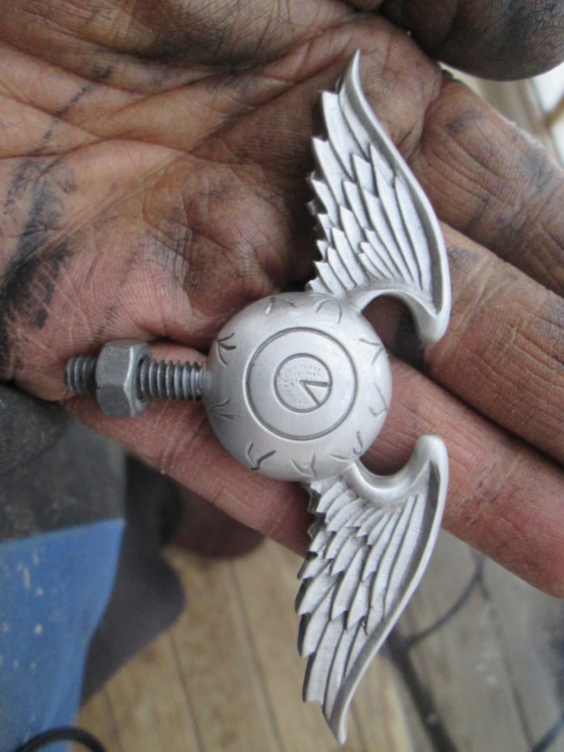 K&ouml;nnte beinhalten: Silberfarbener Metallschmuck mit einem stilisierten Auge mit Fl&uuml;geln. Das Auge hat ein kreisf&ouml;rmiges Design mit einer erhabenen Mitte und einer strukturierten Oberfl&auml;che. Die Fl&uuml;gel sind auf beiden Seiten des Auges ausgebreitet und haben ein detailliertes Federmuster.