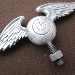 K&ouml;nnte beinhalten: Silbernes Fl&uuml;gelaugen-Ornament mit einer Schraube und einer Mutter zum Anbringen an einem Auto oder Motorrad.