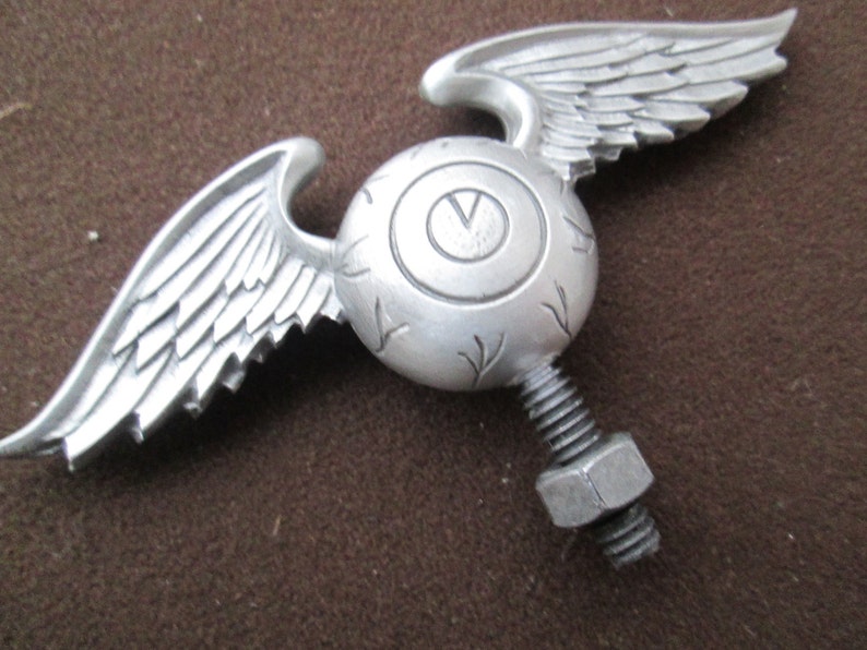 K&ouml;nnte beinhalten: Silbernes Fl&uuml;gelaugen-Ornament mit einer Schraube und einer Mutter zum Anbringen an einem Auto oder Motorrad.