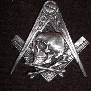 May include: A silver-toned metal pendant featuring a skull and crossbones design within a square and compass, a symbol of Freemasonry.