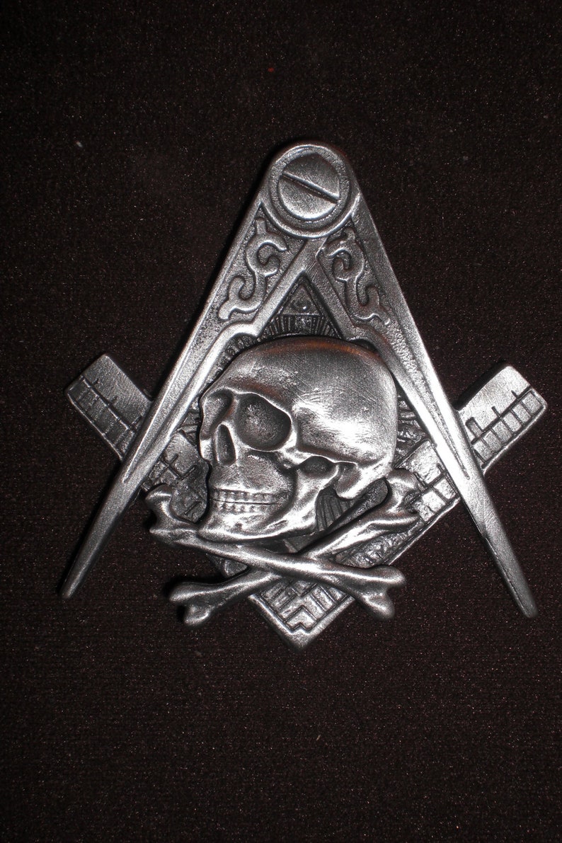 May include: A silver-toned metal pendant featuring a skull and crossbones design within a square and compass, a symbol of Freemasonry.