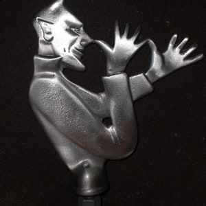May include: A silver-colored metal sculpture of a devil figure with horns, a pointed chin, and outstretched hands. The figure is wearing a coat and is mounted on a bolt. The sculpture has a textured surface.