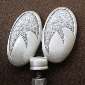 May include: A silver car hood ornament shaped like two stylized ears. The ornament has a textured surface and a threaded base.