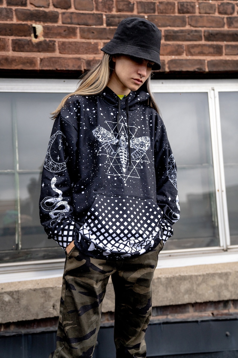 Peut inclure: Un sweat-shirt noir avec un motif g&eacute;om&eacute;trique blanc repr&eacute;sentant un papillon de nuit et des serpents. Le sweat-shirt pr&eacute;sente un motif &agrave; carreaux noir et blanc sur la moiti&eacute; inf&eacute;rieure. Le mod&egrave;le porte un chapeau noir en forme de seau et un pantalon camouflage.