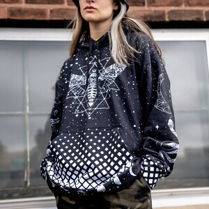 Peut inclure: Un sweat-shirt noir avec un motif blanc imprim&eacute; d'un papillon de nuit, de formes g&eacute;om&eacute;triques et d'&eacute;toiles. Le sweat-shirt a un motif &agrave; carreaux noir et blanc sur la moiti&eacute; inf&eacute;rieure. Le mod&egrave;le porte un chapeau noir en forme de seau.