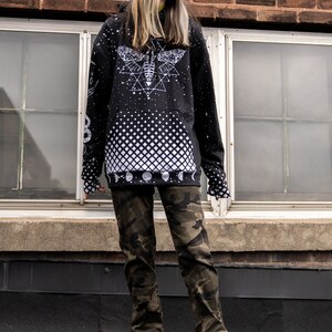 Peut inclure: Un sweat-shirt noir avec un motif blanc imprim&eacute; d'un papillon de nuit et de formes g&eacute;om&eacute;triques. Le sweat-shirt a un motif &agrave; carreaux noir et blanc sur le bas. La personne porte un pantalon camouflage et des sandales noires.