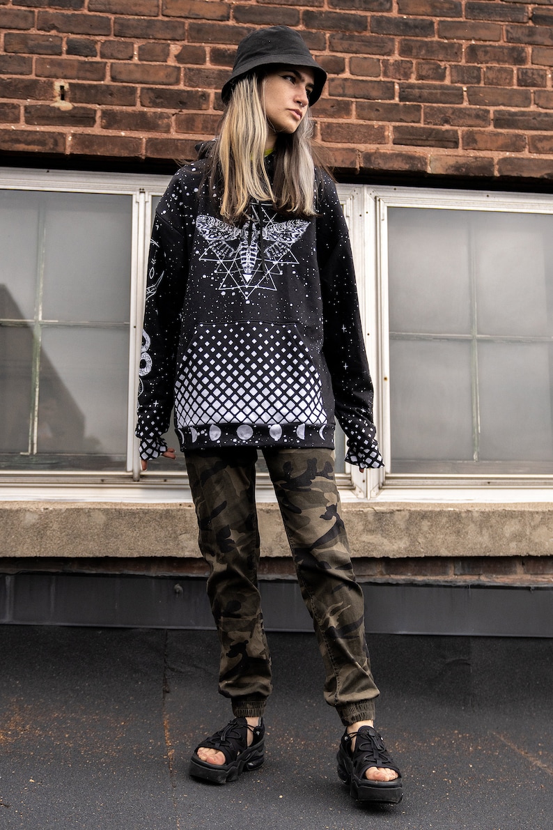 Peut inclure: Un sweat-shirt noir avec un motif blanc imprim&eacute; d'un papillon de nuit et de formes g&eacute;om&eacute;triques. Le sweat-shirt a un motif &agrave; carreaux noir et blanc sur le bas. La personne porte un pantalon camouflage et des sandales noires.