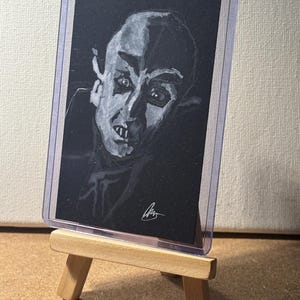Puede incluir: Ilustración en blanco y negro de un vampiro, exhibida en una funda protectora sobre un pequeño caballete de madera. La obra presenta un retrato detallado del rostro del vampiro, con rasgos marcados y colmillos prominentes. La imagen está sobre un fondo oscuro.