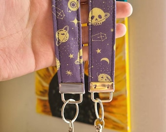 May include: Two purple fabric keychains with gold celestial and skull designs. Each keychain has a silver metal ring and a silver metal clasp.