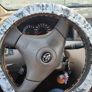 Può includere: Primo piano di un volante di un'auto con un coprivolante a motivi bianco e nero. Il volante ha un rivestimento in pelle nera e un logo Toyota argentato. Il volante è in un'auto con un cruscotto nero.