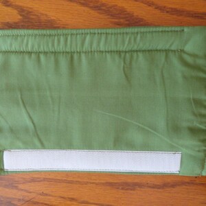 May include: A rectangular, olive green fabric pouch with a white velcro closure. The edges are neatly stitched, and the fabric has a slightly wrinkled texture. The pouch is designed for storage.