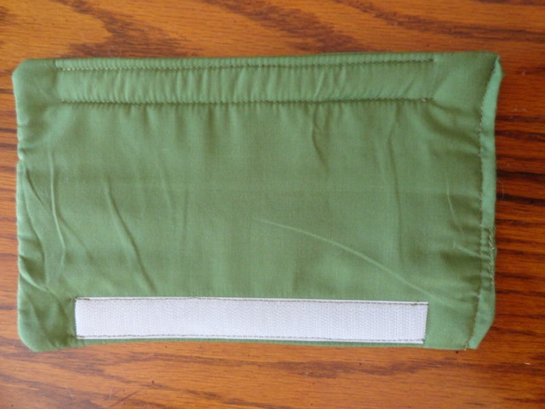 May include: A rectangular, olive green fabric pouch with a white velcro closure. The edges are neatly stitched, and the fabric has a slightly wrinkled texture. The pouch is designed for storage.