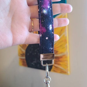 Peut inclure: Un porte-clés en tissu imprimé galaxie noir et rose avec un anneau de clé en métal argenté et un fermoir en métal doré.