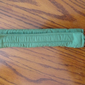 May include: A folded, green fabric item with a series of vertical stitched lines. The item is rectangular and appears to be made of a soft, possibly quilted material. The item is resting on a wooden surface.