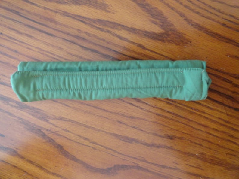 May include: A folded, green fabric item with a series of vertical stitched lines. The item is rectangular and appears to be made of a soft, possibly quilted material. The item is resting on a wooden surface.