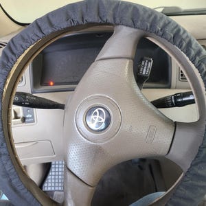 May include: A close-up view of a beige car's steering wheel with a black steering wheel cover. The steering wheel has a silver Toyota logo in the center.