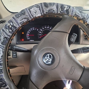 May include: A close-up of a car's steering wheel with a black and white patterned steering wheel cover. The steering wheel is brown and has a silver Toyota logo in the center. The dashboard is visible in the background.
