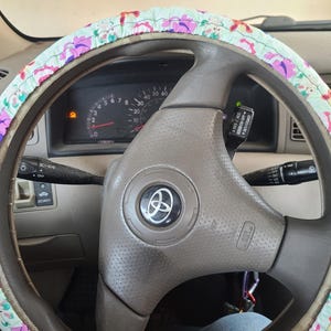 May include: A close-up of a car's steering wheel with a floral patterned steering wheel cover. The steering wheel is brown and has a silver centre with the car's logo. The steering wheel cover is light blue and pink with a floral pattern.