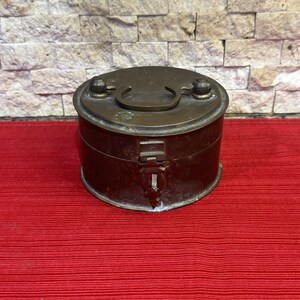 May include: A round, antique brass container with a hinged lid and a handle. The container has a clasp closure and is set against a red background. The brass has a tarnished appearance, suggesting age.