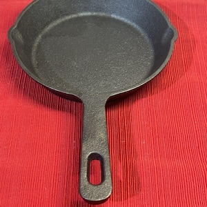 Puede incluir: Una sartén de hierro fundido negra con un solo mango. La sartén está sobre una superficie roja.