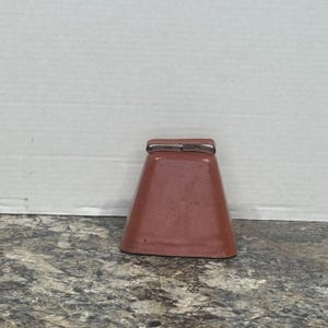 Puede incluir: Una cencerra marrón con una parte superior rectangular y un cuerpo trapezoidal. La campana es de metal y tiene un color marrón oscuro. La cencerra está sobre una superficie texturizada.