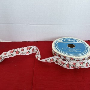 Peut inclure: Une bobine de ruban blanc avec un imprimé floral rouge et vert et une bordure en dentelle blanche. La bobine est étiquetée "Offray Cut Edge Quality Qualite A Bords Coupes Creations Artistiques".