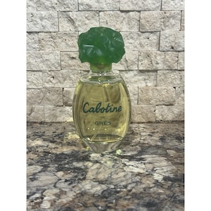 May include: An oval-shaped perfume bottle filled with a light yellow liquid. The bottle has a green, flower-shaped cap. The words "Cabotine de GRES" are written in a cursive font on the front of the bottle. The bottle is sitting on a marble surface.