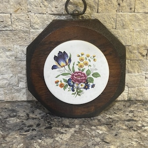 May include: A decorative wall hanging with a dark wood frame and a floral tile insert. The tile features a bouquet of purple, pink, yellow, and blue flowers on a white background. A metal ring is attached at the top for hanging.