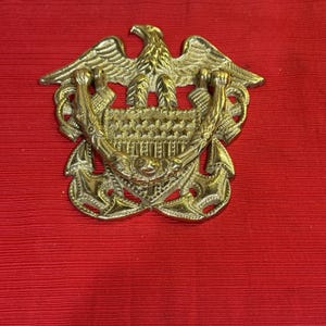Op de afbeelding: Een goudkleurig metalen embleem met een adelaar met uitgespreide vleugels boven een schildontwerp. Het schild wordt geflankeerd door ankers en een decoratieve krans. Het embleem staat tegen een rode achtergrond.