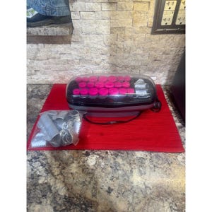 May include: A black and gray electric hair setter with a clear plastic lid containing multiple bright pink hair rollers. Beside the unit is a clear plastic bag filled with various gray plastic hair clips and metal pins.