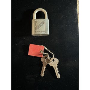 Puede incluir: Un candado plateado con un asa curva y el texto "LAWMAKER" y "RUSTLESS". Dos llaves y una etiqueta roja están unidas al candado con una cadena.
