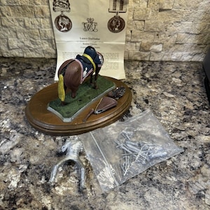 May include: A miniature figurine of a horse and rider, the horse is brown with a yellow mane and tail, the rider is wearing a blue and gold uniform, the figurine is on a wooden base with a green felt surface, the figurine is part of a collection called "All the Queens Men", the text "Lance Daffadar" is visible on the base.