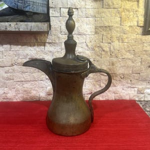 May include: A vintage bronze coffee pot with a long spout and curved handle. The pot has a decorative finial on the lid and is set against a brick wall and a red surface.