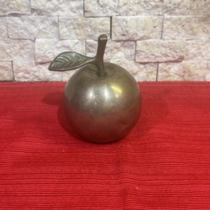 May include: A polished brass apple figurine featuring a smooth, rounded body and a single attached leaf on the stem. The metallic surface reflects light, showing a warm, golden-brown tone throughout the entire structure of the decorative fruit.