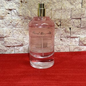 May include: A clear glass perfume bottle with a gold-colored sprayer. The bottle is filled with a pink-tinted liquid and has the words "Floral Magnolia Pour Femme Eau De Parfum" printed on it. The bottle contains 100ml / 3.4 fl oz.